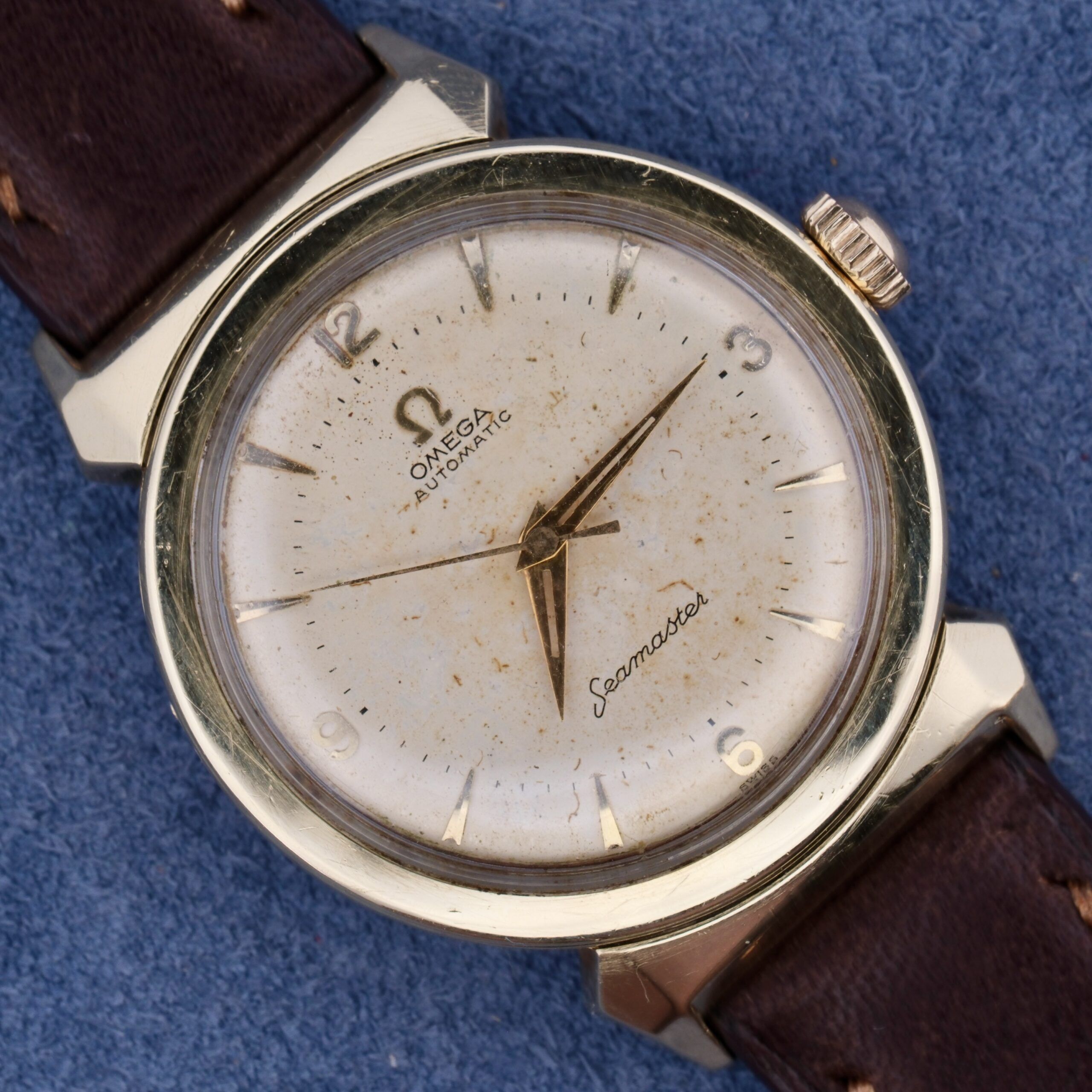 A vintage Omega Semaster with gold-tone casing and bull horn lugs features a white speckled dial, silver markers and hands, and Omega Automatic Seamaster on the face. It has a dark brown leather strap placed on a blue surface.